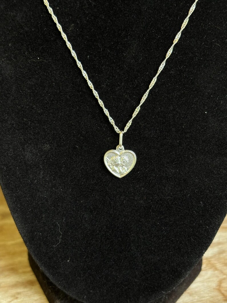 Op de afbeelding: Een zilverkleurige ketting met een hartvormige hanger. De hanger heeft een gedetailleerd reli&euml;fontwerp en hangt aan een delicate ketting. De ketting wordt gepresenteerd tegen een zwarte achtergrond.