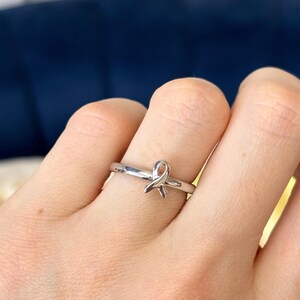 May include: A silver ring with a ribbon design. The ring is on a person's finger.