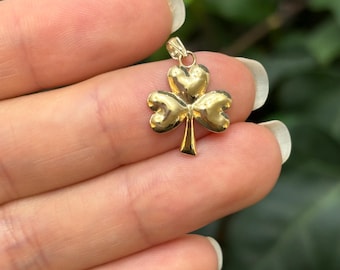 May include: A gold-colored shamrock pendant with three heart-shaped leaves and a stem. The pendant is held up against a green background. The pendant is likely made of gold.