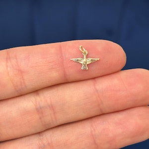 May include: A small, gold-coloured dove charm with outstretched wings. The charm has a loop at the top for attaching to a necklace or bracelet. The dove's body has detailed features, and the charm is held between fingers.