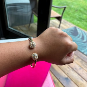May include: A gold-toned open cuff bracelet featuring two spherical ends. Each sphere is encrusted with multiple clear, sparkling stones arranged in a grid pattern. The bracelet has a smooth, polished metal band that curves around the wrist.