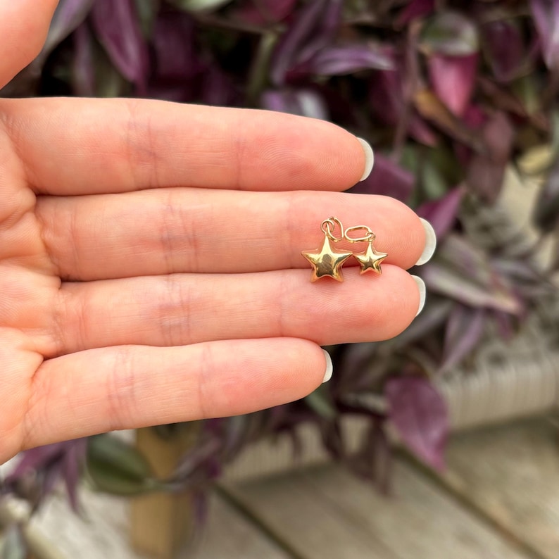 May include: Two gold star charms, one slightly larger than the other, are shown in a hand. The charms are both five-pointed stars.