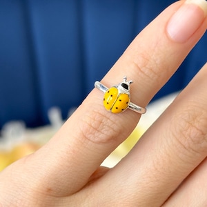 May include: A silver ring with a yellow ladybird design and black spots. The ladybird has a black head and is set on a thin silver band. The ring is a close-up shot, showcasing the details of the insect-themed jewellery.