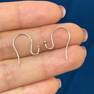 May include: Two gold-colored earring hooks are displayed against a blue background. The hooks have a simple, curved design with a small ball at the bottom of the hook. The hooks are made of a thin, wire-like material.