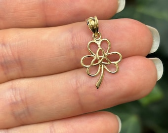 May include: A gold-colored pendant featuring a shamrock design with a Celtic knot in the center. The pendant has a small loop at the top for a chain. The shamrock has three heart-shaped leaves and a stem.