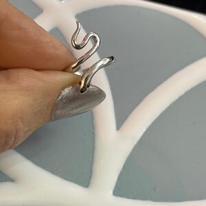 May include: A silver snake ring. The ring's design features a snake wrapping around the finger, with the head and tail pointing upwards. The snake has small red eyes. The ring is held by a person's hand.