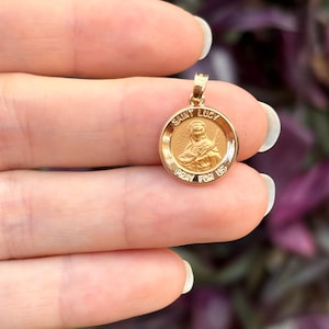 May include: Gold-toned pendant featuring a round medallion with a depiction of Saint Lucy and the text "SAINT LUCY PRAY FOR US".