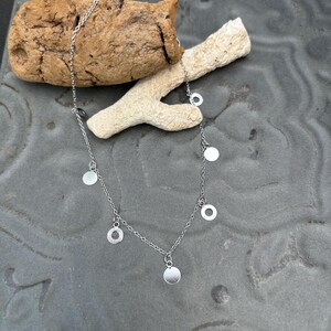 May include: A silver-toned chain necklace featuring multiple small, flat, circular charms spaced evenly along the length of the delicate metal link chain. The necklace is arranged in a curved shape, showcasing the reflective surface of the metal discs.