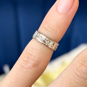 May include: A silver ring with a heart design. The ring is worn on a finger.