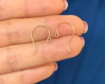May include: Two gold-colored earring hooks are displayed against a blue background. The hooks are curved, with a small loop at the top and a pointed end at the bottom. The earrings are small and delicate.