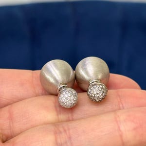May include: A pair of silver-toned stud earrings. Each earring features a large, brushed metal sphere and a smaller, round, crystal-studded sphere. The earrings are displayed against a neutral background.