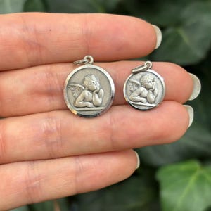 May include: Two silver angel charms, one larger than the other, both depicting a cherub with wings, resting its head on its hand. The charms are oval-shaped and have a raised edge.