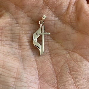 May include: A polished silver-toned metal pendant featuring a cross symbol integrated with a stylized flame design. The pendant includes a small triangular bail at the top for attachment to a chain, showcasing a smooth, reflective metallic finish.