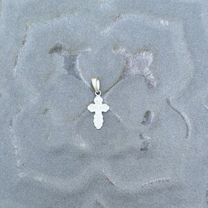 May include: A small silver-toned metal cross pendant featuring an ornate, multi-lobed design with rounded edges and a central cross motif. The pendant includes a simple, smooth metal bail at the top for attachment to a chain or cord.