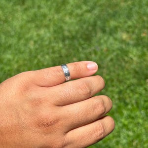 May include: A silver-toned band ring featuring a repeating embossed wave pattern around the exterior surface. The metal ring has a polished finish and a consistent width, showcasing a continuous design of stylized ocean waves encircling the band.