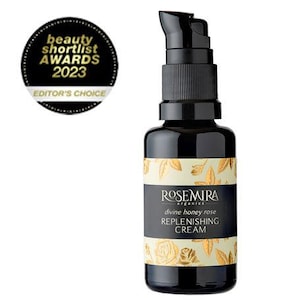 Może przedstawiać: Czarna butelka kremu regenerującego Divine Honey Rose marki Rosemira Organics. Butelka ma złotą etykietę z nazwą produktu i wzorem kwiatowym.