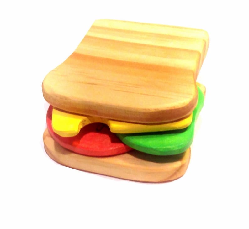 May include: A wooden toy sandwich with two slices of bread, a yellow cheese slice, a red tomato slice, and a green lettuce slice.
