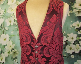 May include: A burgundy vest with a red floral damask pattern. The vest has a V-neck and three silver-colored buttons. The vest is displayed on a mannequin with a floral background.