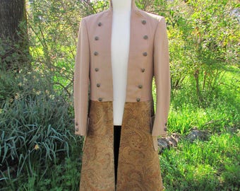 May include: A tan-colored coat with a high collar and multiple buttons. The coat has a patterned, brown fabric panel that extends from the waist down. The coat is displayed on a black mannequin in a natural outdoor setting.