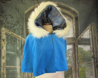 May include: A blue cape with a white faux fur trimmed hood. The hood's interior is a patterned blue fabric. The cape has a zipper closure and is displayed on a mannequin. The background features a distressed window.