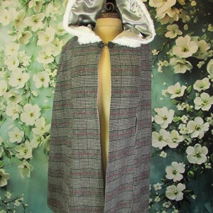 May include: A gray plaid cape with a hood lined in silver satin and trimmed with white faux fur. The cape is fastened with a decorative clasp and displayed on a mannequin. The background features a floral pattern.