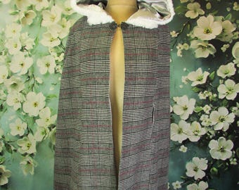 May include: A gray plaid cape with a hood lined in silver satin and trimmed with white faux fur. The cape is fastened with a decorative clasp and displayed on a mannequin. The background features a floral pattern.