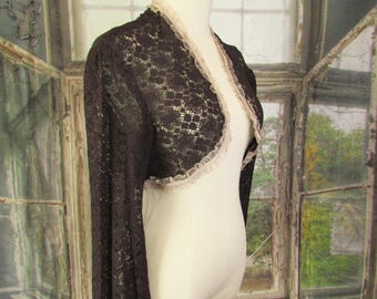 May include: A dark brown lace bolero jacket with long sleeves and a cream-colored trim. The jacket is displayed on a mannequin, showcasing its delicate floral pattern and elegant design. The bolero adds a touch of vintage charm.
