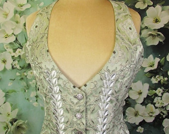 May include: A light green vest with a halter neckline and a decorative floral pattern. The vest has silver-colored leaf-like embroidery and decorative buttons down the front. The vest is displayed on a mannequin.