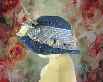May include: A blue floral print hat with a white and blue plaid ribbon and a white lace trim. The hat has a rounded crown and a wide brim. A white flower is attached to the side of the hat. The hat is displayed on a mannequin head.