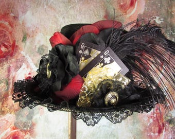 May include: A black top hat with a red velvet trim and a black feather. The hat is decorated with a black rose, gold fabric, and two playing cards: the Queen of Spades and the Queen of Clubs.