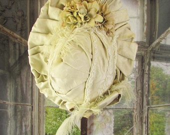 May include: Cream-colored vintage hat with a wide brim and floral embellishments. The hat features a ruffled design, lace trim, and delicate feathers. It is displayed on a brown metal stand against a backdrop of a window with a blurred outdoor scene.