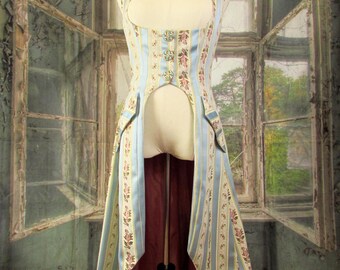 May include: A light blue and cream-colored vest with vertical stripes and floral patterns. The vest has a high collar, decorative buttons, and a long, flowing back. The lining is a deep red color. The garment is displayed on a mannequin.