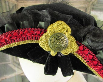 May include: A black hat with red and gold trim, and a gold decorative button. The hat has a velvet texture and is adorned with black ribbon and ruffled fabric. The hat is likely a vintage or costume piece.