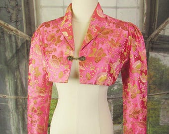 May include: A pink bolero jacket with a floral pattern in gold. The jacket has long sleeves and a decorative clasp closure. The garment is displayed on a mannequin.