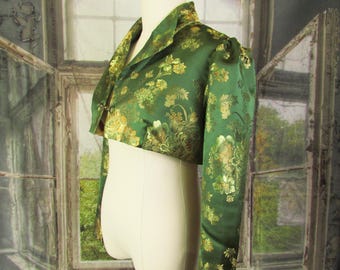 May include: A cropped, emerald green jacket with long sleeves and a mandarin collar. The jacket is adorned with a gold floral pattern. The garment is displayed on a mannequin, showcasing its tailored fit and design.