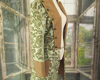 May include: A long, olive green and tan coat with a floral pattern, displayed on a mannequin. The coat features a tan collar and lapels, with decorative buttons and a chain detail. The coat's design is reminiscent of historical or theatrical attire.