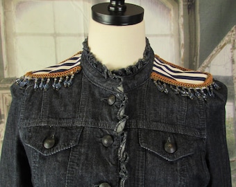May include: Dark denim jacket with decorative shoulder accents. The jacket features a high collar, button-up front, and ruffled trim. Shoulder details include striped ribbon, gold trim, and beaded fringe. The jacket has two front pockets and dark buttons.