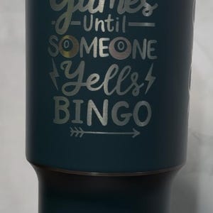 May include: A teal tumbler with a silver inscription that reads "It's All Fun & Games Until Someone Yells Bingo" with a silver arrow pointing to the right. The inscription includes three silver bingo balls with the numbers 08, 12, and 42.
