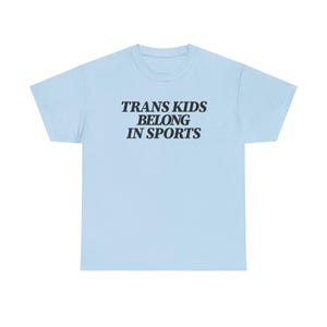以下が含まれることがあります： 胸の中央に太字のサンセリフ体で「TRANS KIDS BELONG IN SPORTS」という黒いテキストがプリントされた、ライトブルーの半袖Tシャツ。クルーネックで、標準的でゆったりとしたフィット感の衣類です。
