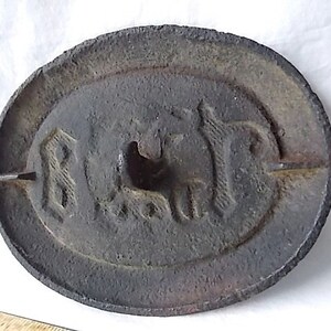 May include: A circular, cast iron weight with an oval central design. The weight has embossed text and a horse image. The dark grey metal has a rough, textured surface, showing signs of age and use.