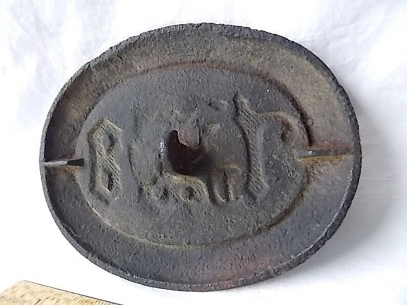 May include: A circular, cast iron weight with an oval central design. The weight has embossed text and a horse image. The dark grey metal has a rough, textured surface, showing signs of age and use.