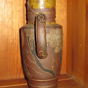 Peut inclure: Une cruche en c&eacute;ramique brune avec une bande jaune moutarde sur le haut orn&eacute;e de petites &eacute;toiles et de motifs floraux. Le corps pr&eacute;sente des paysages grav&eacute;s aux contours dor&eacute;s, incluant des arbres et une anse avec des volutes dor&eacute;es.
