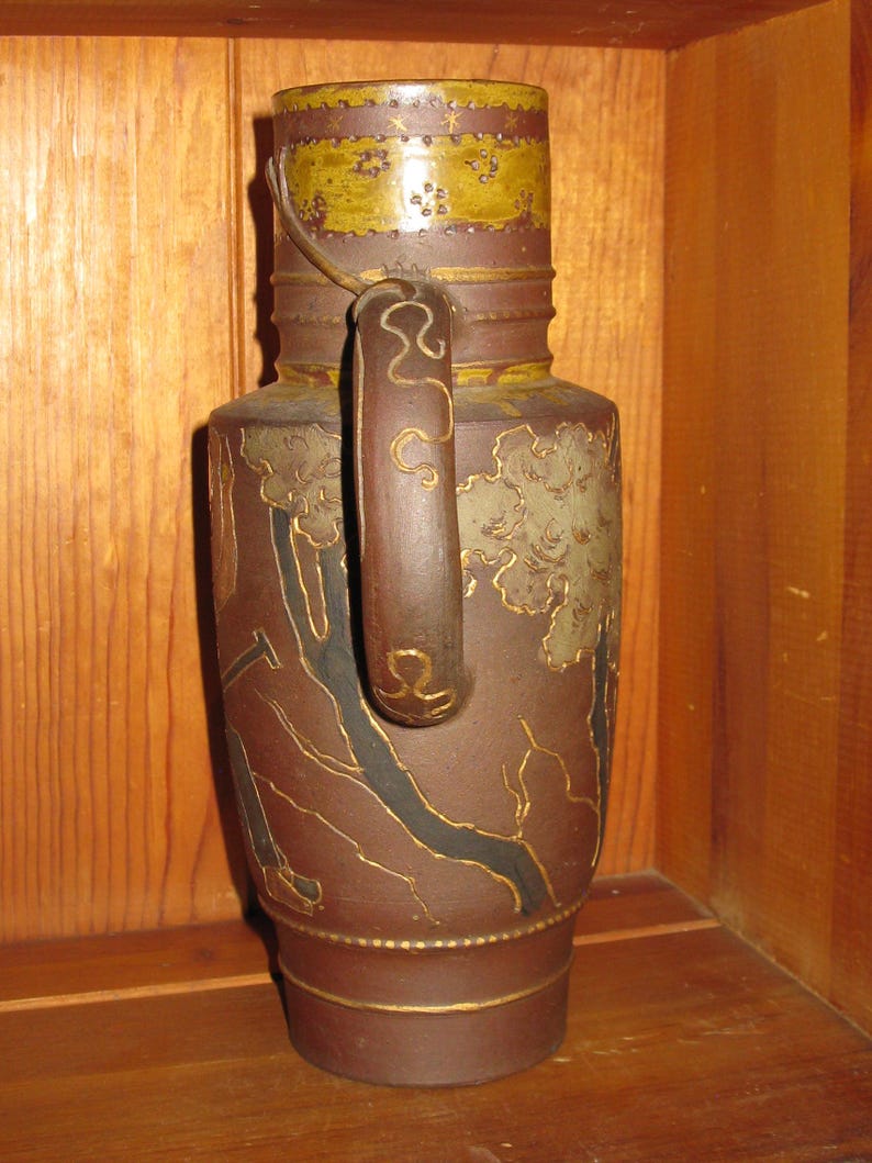 Peut inclure: Une cruche en c&eacute;ramique brune avec une bande jaune moutarde sur le haut orn&eacute;e de petites &eacute;toiles et de motifs floraux. Le corps pr&eacute;sente des paysages grav&eacute;s aux contours dor&eacute;s, incluant des arbres et une anse avec des volutes dor&eacute;es.