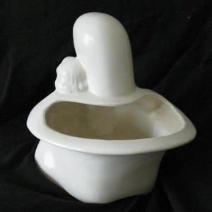 May include: A white ceramic planter in the shape of a mother and child. The planter has a rounded base and a wide, shallow opening. The mother figure is tall and slender, with the child nestled against her. The planter is on a black background.