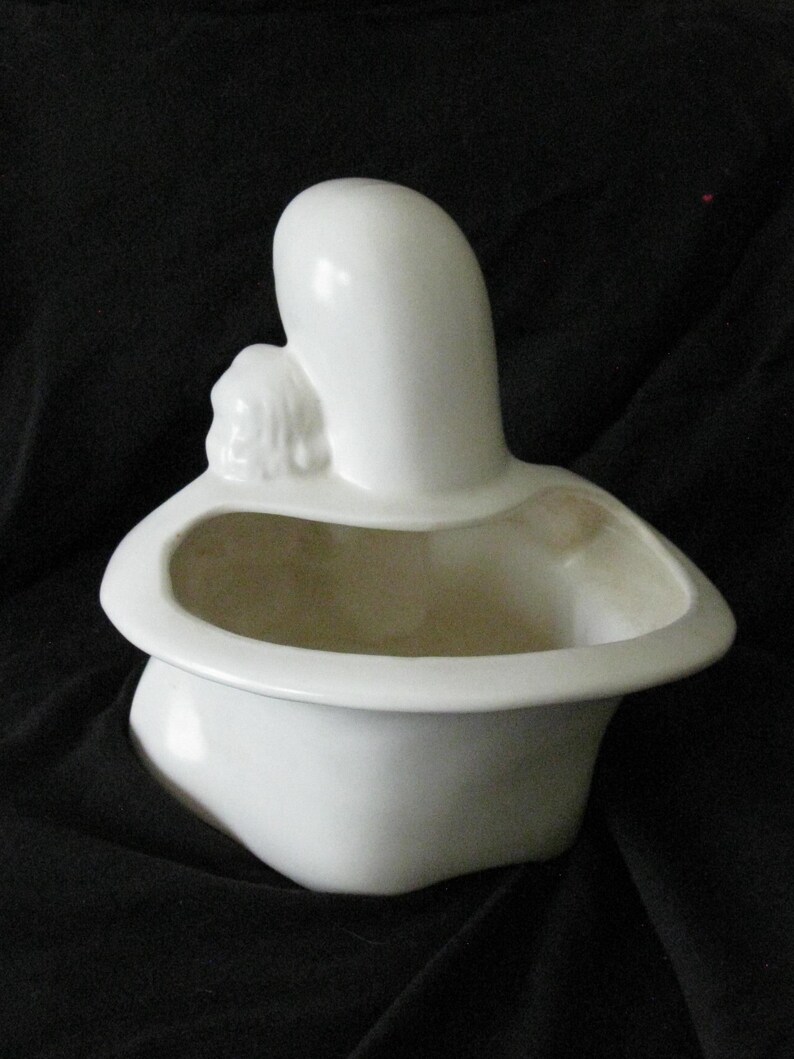 May include: A white ceramic planter in the shape of a mother and child. The planter has a rounded base and a wide, shallow opening. The mother figure is tall and slender, with the child nestled against her. The planter is on a black background.