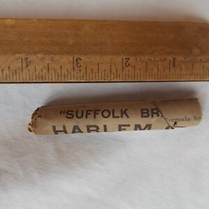 May include: A vintage wooden ruler with inch markings next to a small, cylindrical object wrapped in tan paper. The paper is printed with the words "SUFFOLK BR" and "HARLEM". The ruler is approximately 5 inches long.