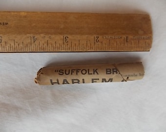 May include: A small, cylindrical, light brown paper wrapper with black text reading "SUFFOLK BR" and "HARLEM". The wrapper is positioned below a wooden ruler that shows a length of approximately 2.5 inches for the paper object.