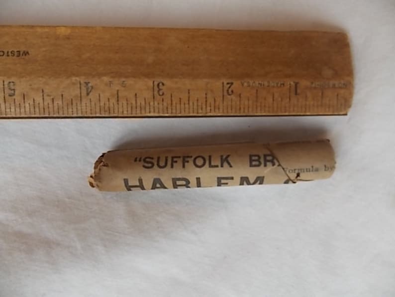 May include: A vintage wooden ruler with inch markings next to a small, cylindrical object wrapped in tan paper. The paper is printed with the words "SUFFOLK BR" and "HARLEM". The ruler is approximately 5 inches long.
