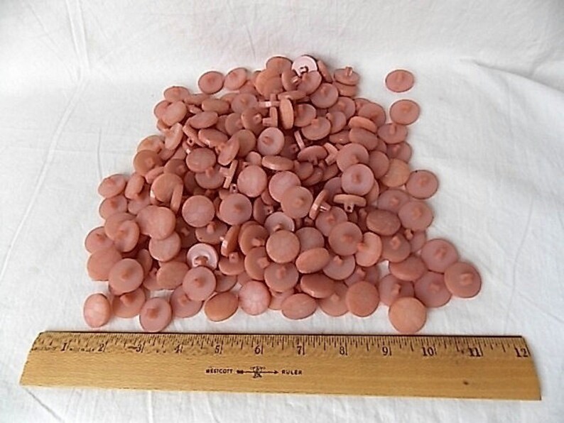May include: A pile of light pink buttons with a flat, round design. A wooden ruler with inch markings is placed at the bottom of the buttons. The buttons are scattered on a white surface.