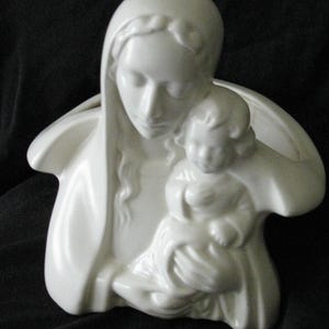 May include: A white ceramic sculpture depicting a mother holding a child. The figures are draped in flowing garments, with the mother's head covered by a veil. The sculpture has a smooth, glossy finish and is set against a dark background.